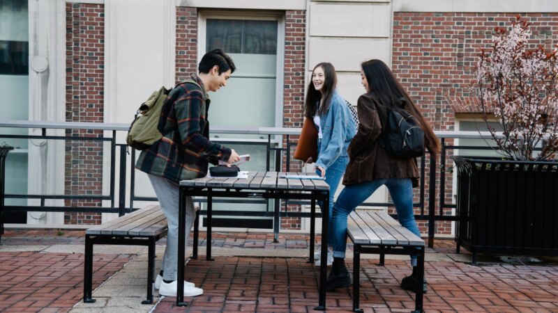 Two students sitting on a campus bench, one looking at their phone, the other gazing away